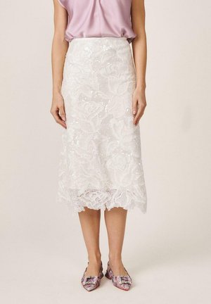 Mujer con falda midi blanca de encaje con patrones florales, blusa rosa sin mangas y zapatos planos puntiagudos multicolores con adornos.