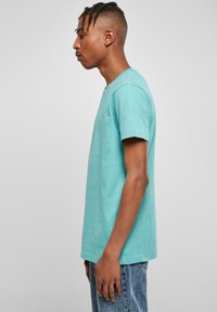 Tricou din bumbac turcoaz, cu guler clasic și mâneci scurte, prezentat pe o persoană întoarsă de profil. Designul este simplu și neîmpodobit.