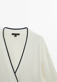 V-neck cardigan in cream with navy trim. Features a ribbed texture, wide sleeves, and a label at the neckline.