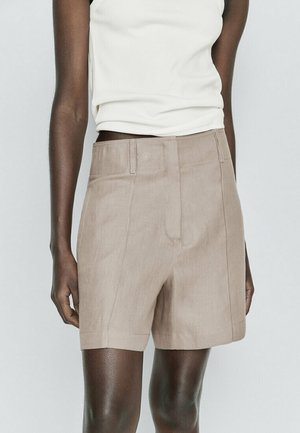 Henkilö, jolla on yllään beige korkeavyötäröiset räätälöidyt shortsit ja hihaton valkoinen ribbipaita, kohoaa vaaleaa taustaa vasten.