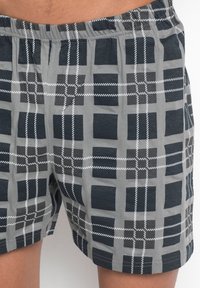 Un short à carreaux gris avec un motif géométrique comportant des carrés bleu foncé et des lignes blanches. Taille élastique, tissu léger, design décontracté.