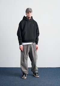 Sort hættetrøje, grå stribede baggy bukser og sorte sneakers med hvide og beige detaljer, afsluttet med en grå kasket.