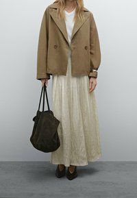 Frau trägt eine beigefarbene, cropped Jacke, einen hellen, plissierten Maxirock, schwarze, spitze Schuhe und hält eine große, dunkelbraune Tragetasche.