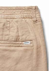Beige pamut nadrág textúrált felülettel, hátul zsebbel és egy fehér címkével, ami a "GABBA" feliratot viseli. Összevarrási részletek a derekán.