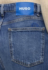 Vue arrière d'un jean en denim bleu montrant une grande poche arrière et un patch de marque en cuir bleu avec "HUGO" en lettres blanches.