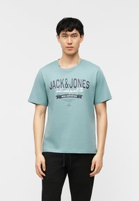 Jeune homme debout, tourné vers l'avant, portant un t-shirt Jack & Jones bleu sarcelle clair et un pantalon noir sur un fond blanc uni.