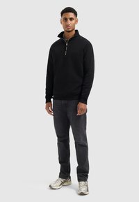 Zwarte zip-up sweatvest met hoge kraag, zachte stof, geribde manchetten, gecombineerd met donkere jeans en metallic sneakers.