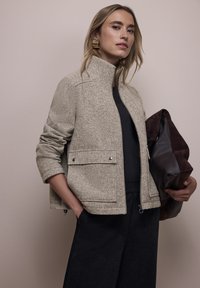Hellbeige Tweedjacke mit Stehkragen, silbernen Knöpfen und zwei vorderen Taschen; kombiniert mit dunklen, weit geschnittenen Hosen und einer braunen Tasche.