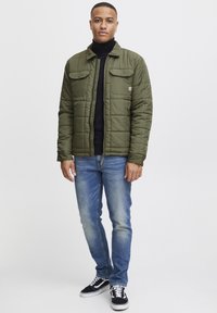 Gequilted olijfgroene jas met twee borstzakken, gecombineerd met een zwarte turtleneck, blauwe spijkerbroek en zwarte sneakers. Gladde textuur, casual pasvorm.