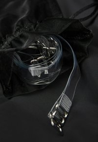 Genomskinlig förvaringsbehållare med silverfärgade metallklämmor och genomskinliga remmar, placerad på en svart textilbakgrund. Remmarna har texturerade ytor.