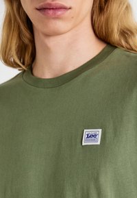 T-shirt in cotone verde oliva con collo rotondo e piccola patch logo blu e bianco di Lee Workwear sul lato sinistro. Texture liscia.
