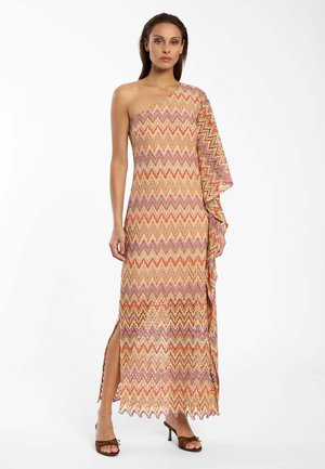 Frau trägt ein einseitig schulterfreies, knöchellanges Kleid mit Zickzackmuster in Orange-, Pink- und Beigetönen, kombiniert mit braunen Sandalen mit Absatz.