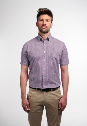 Kurzarmhemd aus einem gemusterten Stoff mit roten und blauen geometrischen Formen, mit button-down Kragen und einer Brusttasche. Getragen mit beigen Hosen.