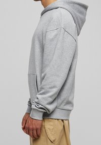 Felpa grigia in cotone con tasca frontale, polsini a costine e cappuccio con coulisse. Texture liscia e vestibilità rilassata, indossata con pantaloni corti beige.