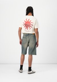 Camiseta beige con gráfico de sol rojo y texto, combinada con pantalones cortos de denim azul. El conjunto se complementa con calcetines negros y zapatillas deportivas blancas.