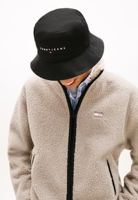 Μαύρο καπέλο τύπου bucket με το λογότυπο "TOMMY JEANS". Ανοιχτό μπεζ μπουφάν από φλις με φερμουάρ μπροστά και πλαϊνές τσέπες. Συνδυασμένο με πουκάμισο με μπλε ρίγες.