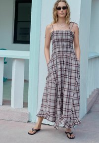Vestido maxi con estampado de cuadros que presenta tirantes ajustables, un corsé elástico fruncido, bolsillos laterales y un dobladillo con volantes. Se combina con sandalias negras.