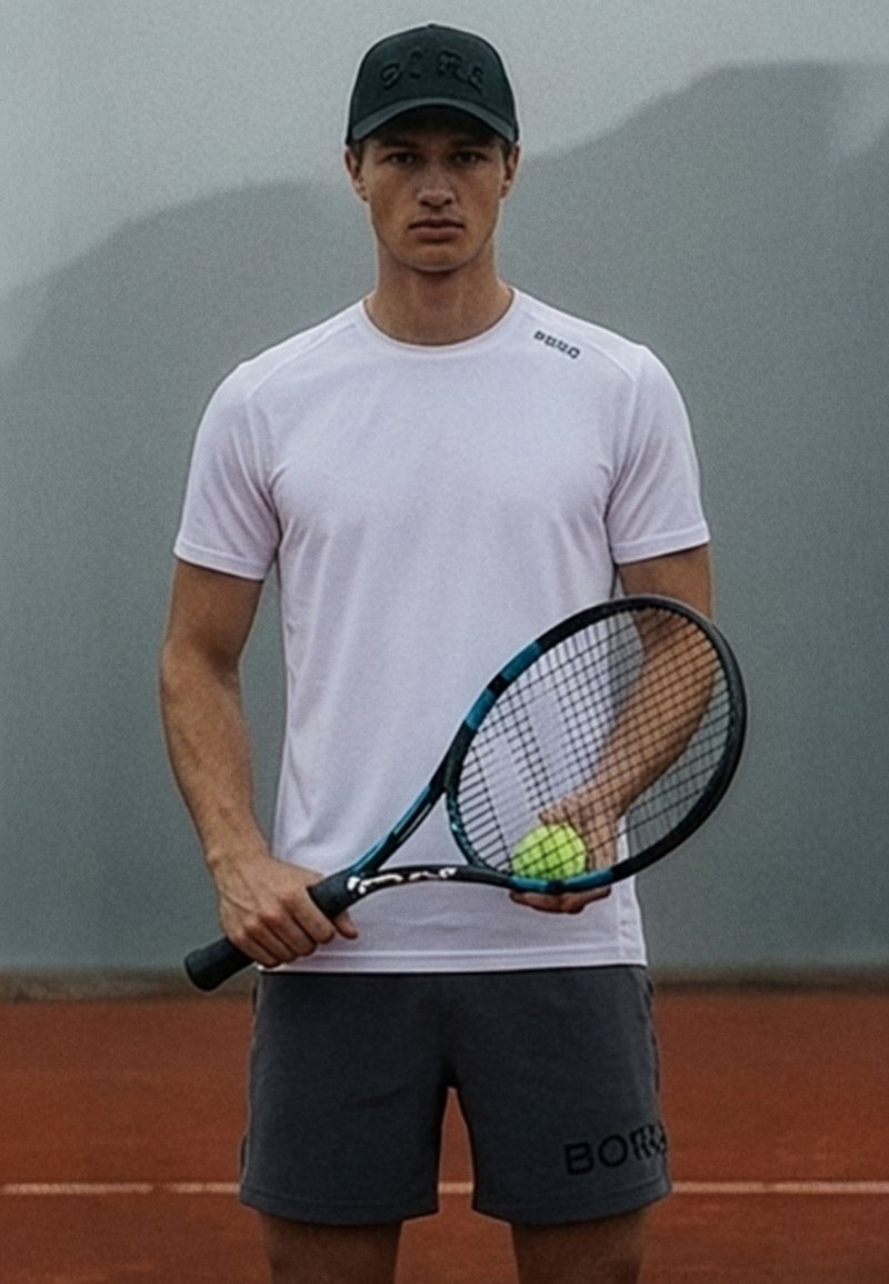 Fiatal férfi sportfelszerelésben teniszüstököt tart a kezében egy salakos teniszpályán felhős ég alatt.