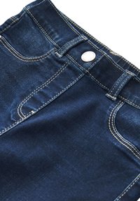 Gros plan sur un jean en denim bleu foncé montrant la poche avant, les passants de ceinture et le bouton métallique avec des coutures contrastantes blanches.