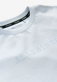 Světle modré bavlněné tričko s kulatým výstřihem, zdobené vyšívaným logem ve stejné barvě a kontrastní černou štítkou uvnitř límce.