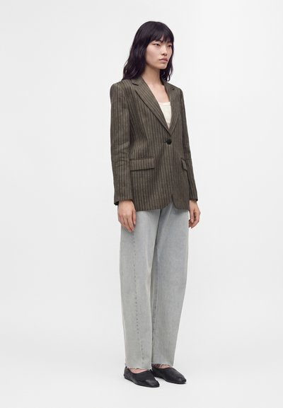 Femme aux longs cheveux noirs portant un blazer marron à fines rayures, un jean large gris clair, des chaussures plates noires et un haut beige, debout sur fond blanc.