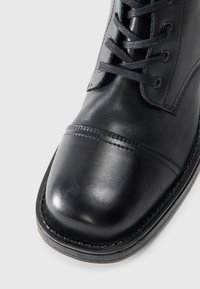Schwarze Lederstiefel mit abgerundetem Zehenbereich, glatter Textur, gesteppten Details und schwarzen Schnürsenkeln. Verfügt über eine glänzende Oberfläche und eine robuste Sohle.