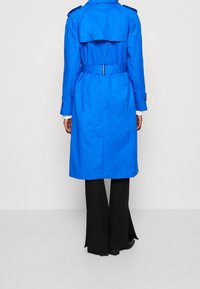 Niebieski trencz z paskiem w talii, detalem z podwójnym rzędem guzików i pagonami na ramionach. Czarne, rozszerzane spodnie częściowo widoczne poniżej.