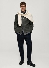 Grijze wollen jas met ritssluiting, twee frontzakken; gedragen over een donker shirt. Marineblauwe broek en zwarte schoenen maken de outfit compleet. Sjaal hangt over de schouders.