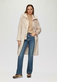 Manteau long en tartan beige sur un pull beige, associé à un jean évasé bleu et des baskets marron. Textures douces, coupe décontractée, look superposé.