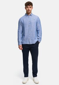 Jonge man die staat met één hand in zijn zak, draagt een lichtblauw overhemd met knopen, donkere broek en witte sneakers tegen een witte achtergrond.