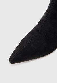 Scarpa nera in suede con tacco alto e punta affusolata, caratterizzata da una superficie liscia e cuciture visibili lungo le cuciture.