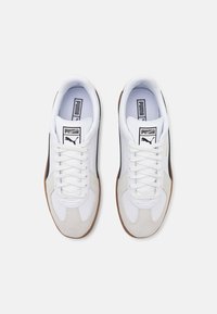 Zapatilla blanca y gris con un upper de ante y cuero, que presenta acentos negros, cordones blancos y una suela de goma color caramelo. Logotipo de Puma en la lengua.