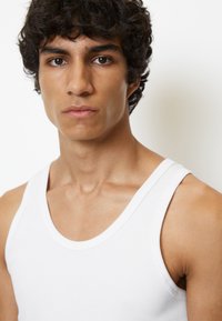 Top sin mangas de algodón blanco con cuello redondo; el ajuste ceñido resalta los hombros y la clavícula. El fondo es de un color claro uniforme.