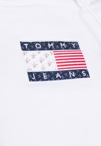 Bílá mikina s vyšívanou obdélníkovou nášivkou s červenými a modrými pruhy, hvězdami a textem "TOMMY JEANS" v námořnické modři.