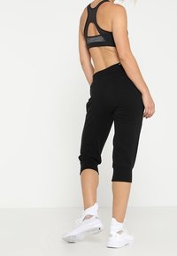 Mujer vestida con un sujetador deportivo negro, pantalones de chándal cortos negros y zapatillas deportivas blancas, de pie contra un fondo blanco con la espalda vuelta.