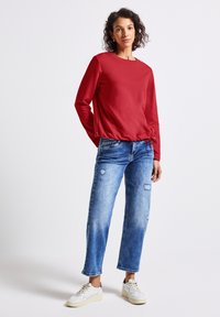 Czerwona bluza z długim rękawem, okrągłym dekoltem i sznurkiem do regulacji u dołu, zestawiona z przetartymi niebieskimi dżinsami i białymi sneakersami.