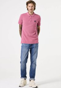 Camisa polo rosa hecha de algodón con cuello, mangas cortas y un logo en el pecho. Combinada con jeans azul claro y zapatillas beige.