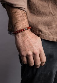 Brățară din mărgele roșii din lemn, cu un accent metalic, odihnindu-se pe o mână. Mâna este parțial strânsă și are fire de păr deschise la culoare.