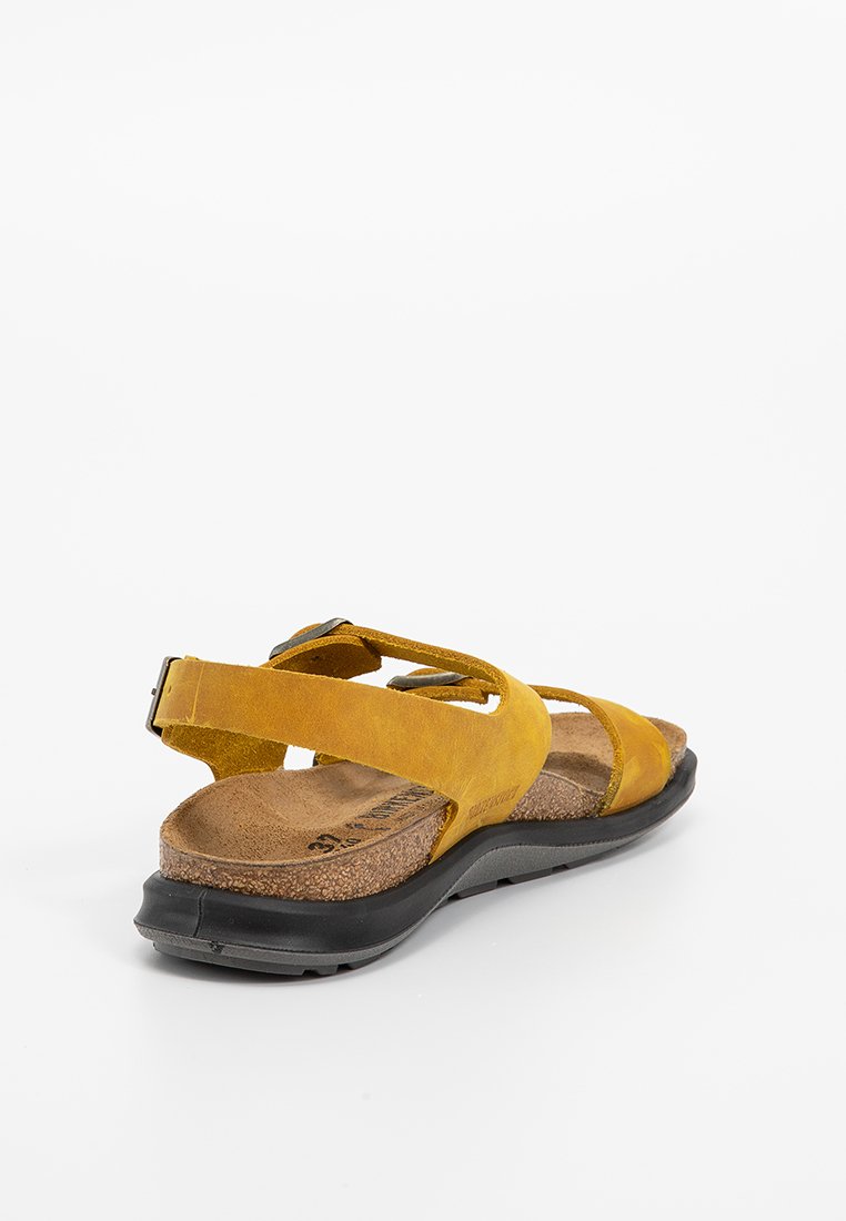 Sandale en suede jaune avec des lanières entrecroisées, semelle en liège et semelle en caoutchouc noir. Dispose d'une sangle de cheville ajustable pour un bon maintien.