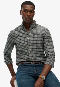 Camisa de cuadros en tonos azul marino y crema con botones y mangas largas, combinada con jeans de color azul oscuro y un reloj de cuero marrón. Cuenta con un bolsillo en el pecho y botones.