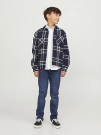 Πουκάμισο με καρό σε ναυτικό μπλε και λευκό πάνω από ένα λευκό πουκάμισο με κουμπιά, συνδυασμένο με σκουρό μπλε τζιν και μαύρα αθλητικά παπούτσια, με άνετη εφαρμογή και casual στυλ.