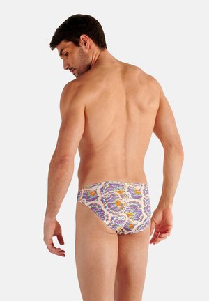 Man standing turned back, wearing colorful floral-patterned swim briefs against a plain white background.