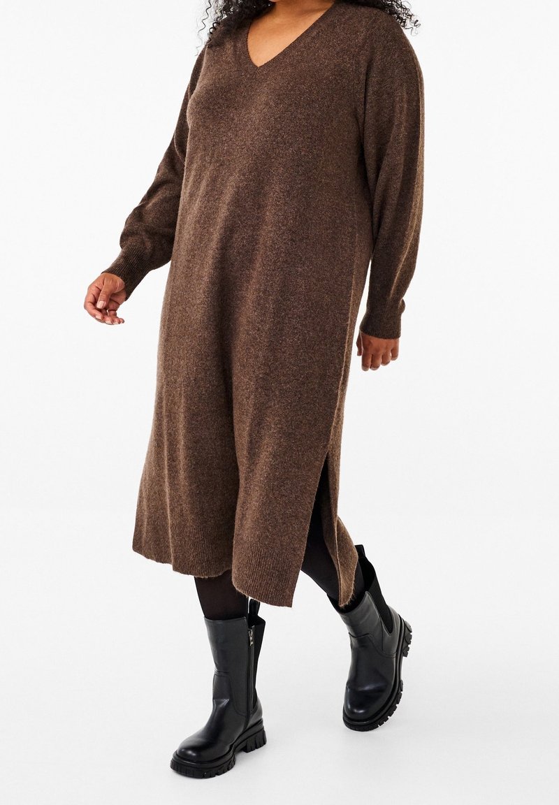 Femme portant une robe-pull longue marron à col en V avec des fentes sur les côtés, des collants noirs et des bottes en cuir noires épaisses, marchant vers l'avant.