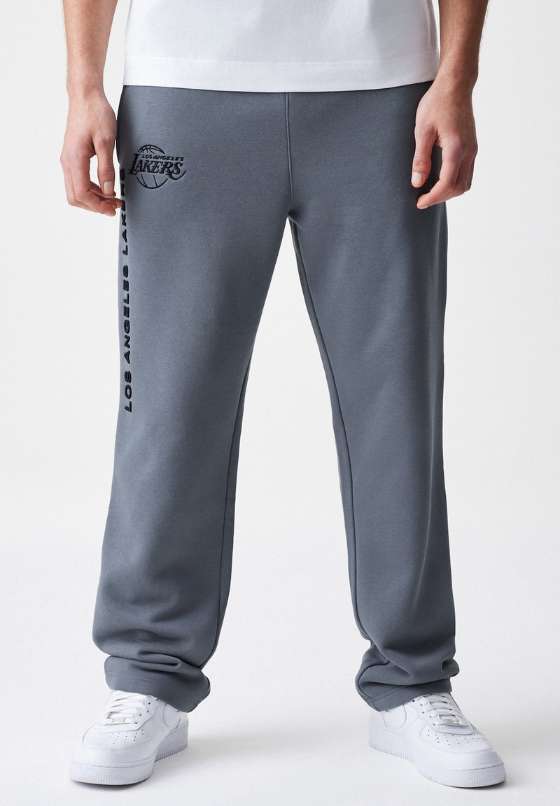 Graue Fleece-Sweatpants mit lockerer Passform, versehen mit einem Logo der "Los Angeles Lakers" und vertikalem Text entlang des linken Beins. Akzentuiert mit weißen Sneakers.