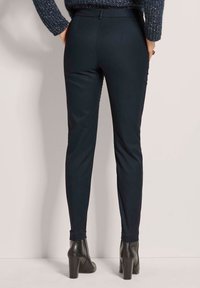 Marineblaue, taillierte Hose mit glatter Textur, maßgeschneidertem Design und cropped Beinen. Kombiniert mit schwarzen High Heels.