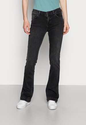 Person iført sorte bootcut-jeans, hvide sneakers og en turkis trøje, der står mod en ensartet lysegrå baggrund.