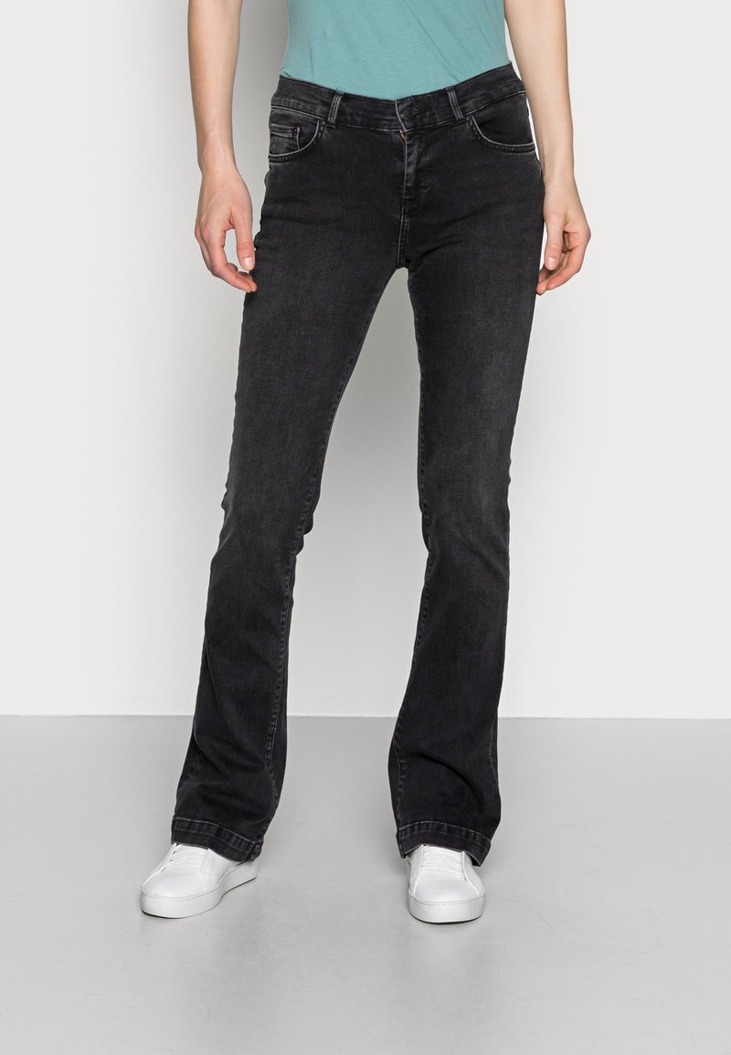 Personne portant un jean bootcut noir, des baskets blanches et une chemise turquoise, debout devant un fond uni gris clair.