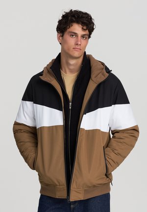 Giovane uomo che indossa una giacca a zip color block marrone, bianca e nera con le mani nelle tasche, in piedi contro uno sfondo neutro.