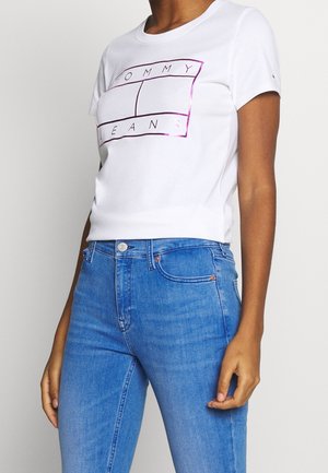 Osoba ma na sobie białą koszulkę z krótkim rękawem z fioletowym logo "Tommy Jeans" oraz niebieskie jeansy z wysokim stanem.