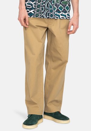 Pantaloni in cotone beige dalla vestibilità dritta, con tasche laterali, abbinati a scarpe in suede verde. Lo sfondo è bianco uniforme.